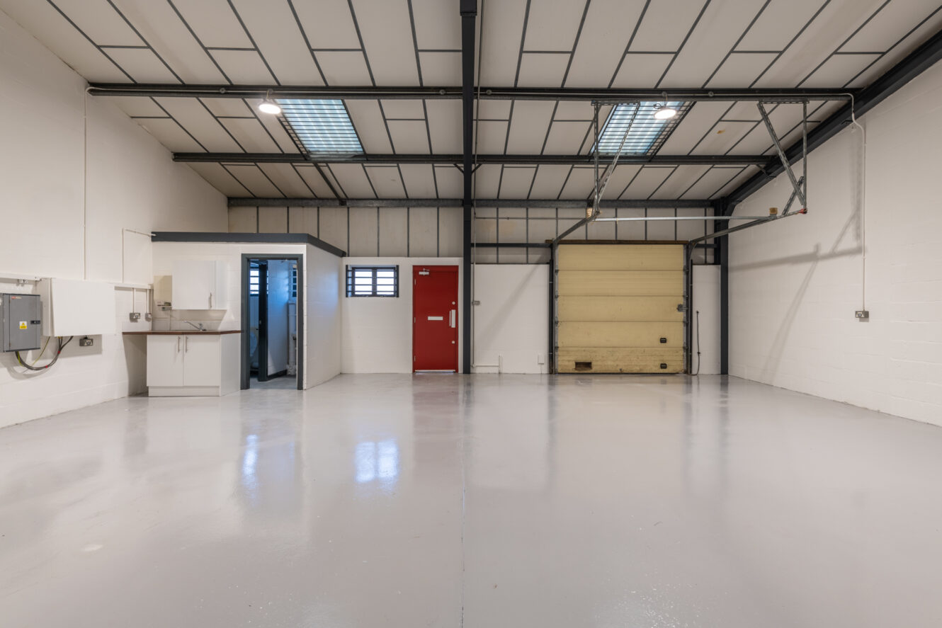 Empty industrial warehouse unit with white walls, grey floor, overhead lights, a red door, a beige roller shutter door, sink, and small restroom area on the left.