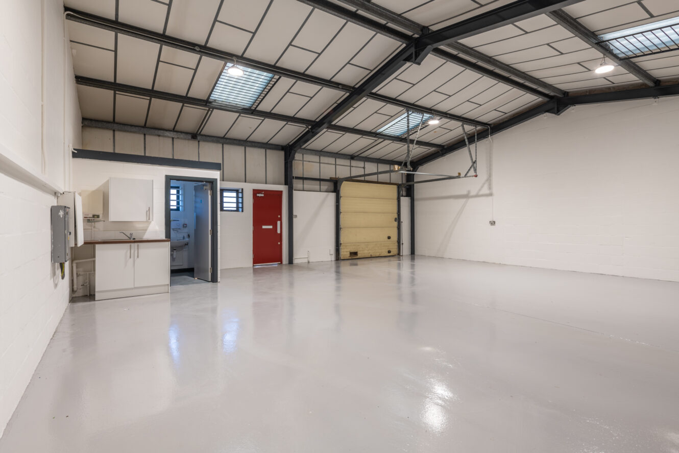 Clean, empty warehouse interior with glossy concrete floor, high ceiling, skylights, a red door, a yellow garage door, and a small kitchenette area on the left.