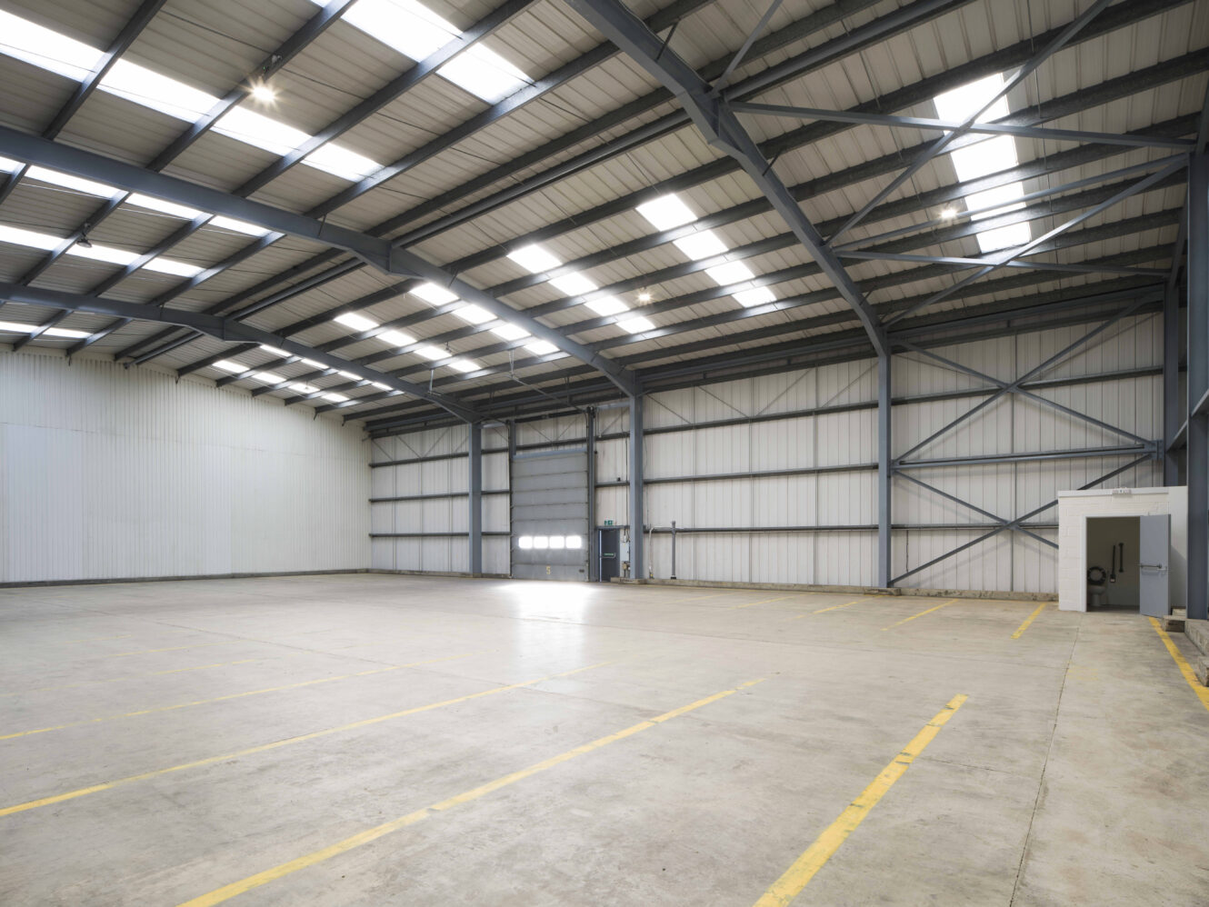 Interior of an empty, clean warehouse with high ceilings, exposed steel beams, skylights, and marked parking spaces on the concrete floor.