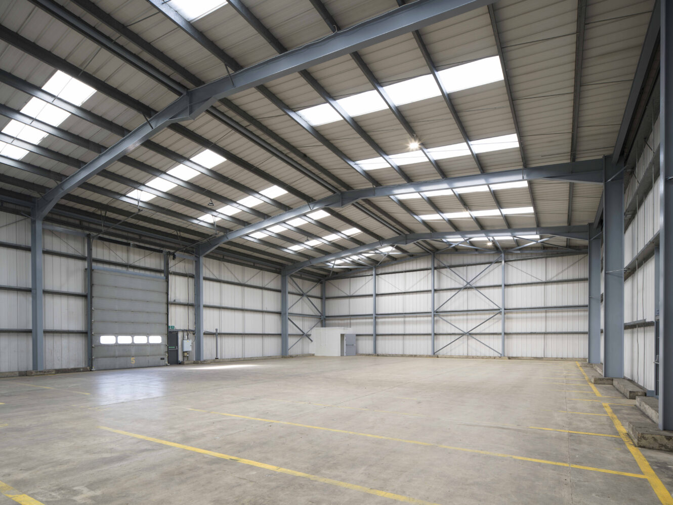 Interior of an empty warehouse with a high ceiling, exposed steel beams, concrete floor, and large overhead doors. Yellow lines mark the floor for vehicle or storage bays.