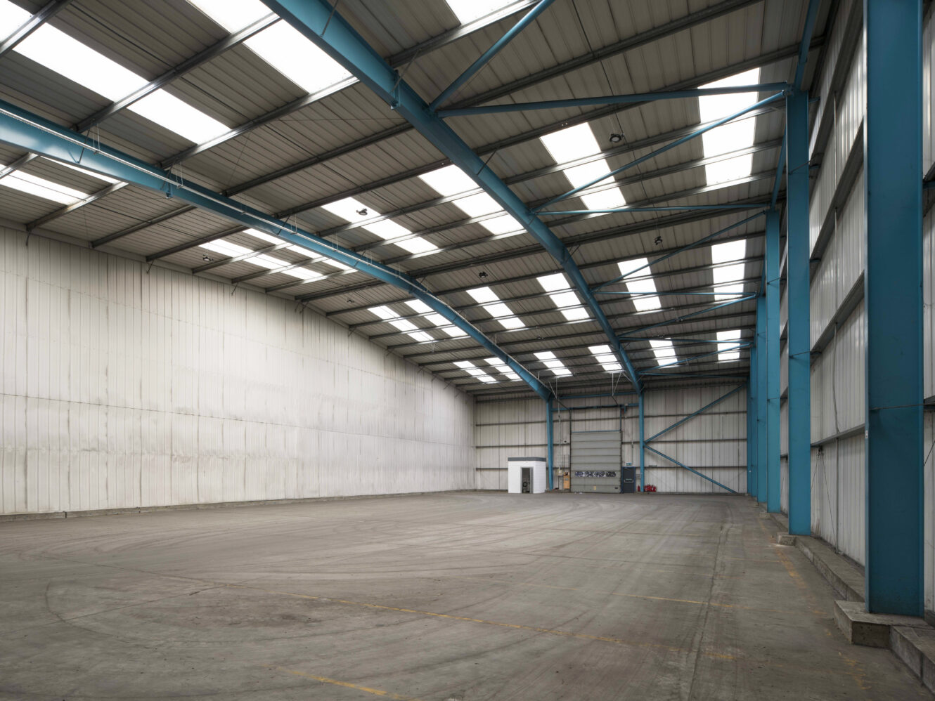 Spacious, empty industrial warehouse with high ceilings, blue steel beams, skylights, and a small white office in the corner.