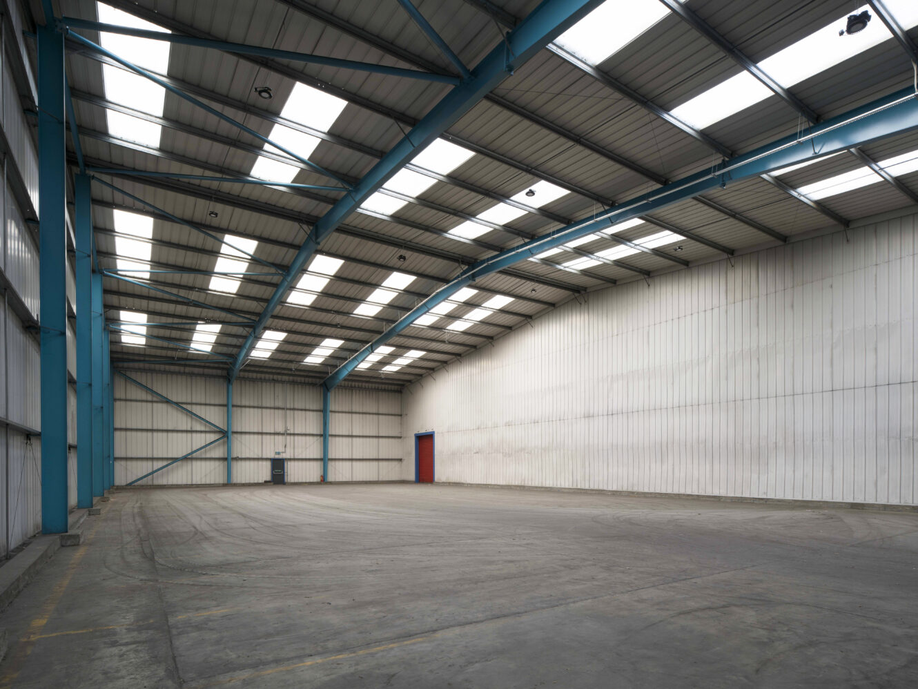 Large empty warehouse with high ceilings, skylights, white paneled walls, concrete floor, and a single red door on the far wall.