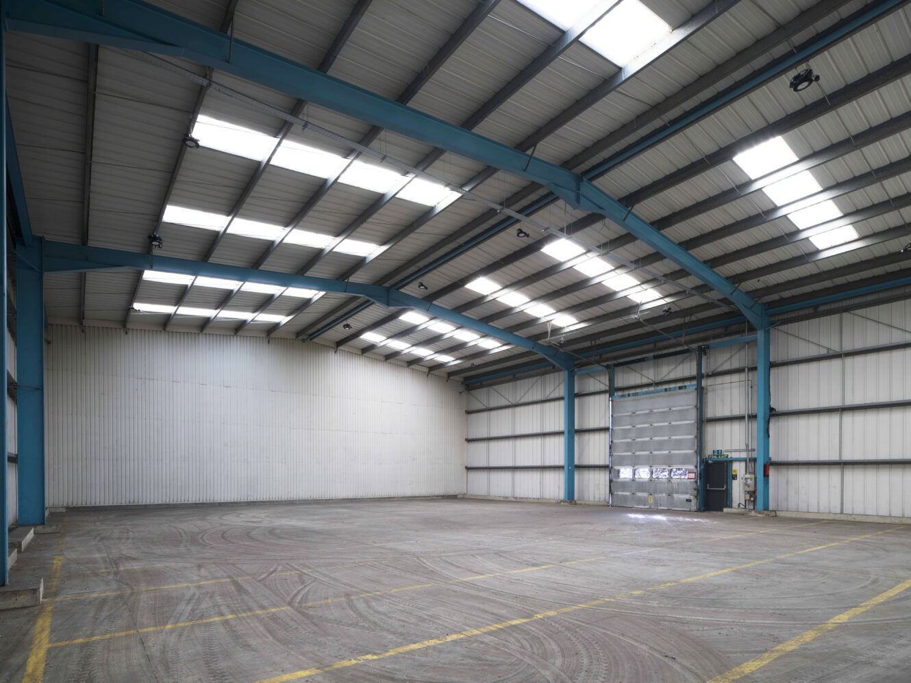 Interior of a large, empty industrial warehouse with high ceilings, blue steel beams, skylights, and a closed roller shutter door.