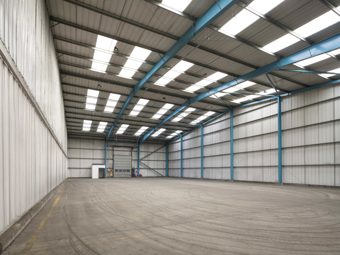 Empty industrial warehouse with high ceilings, skylights, blue steel beams, and white corrugated metal walls. The concrete floor shows faint tire marks.