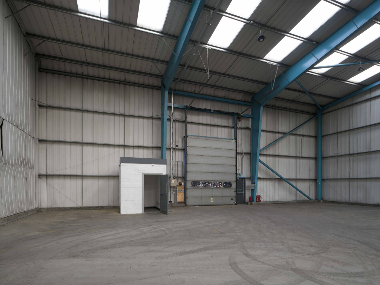 Empty industrial warehouse interior with high ceilings, metal walls, a large roller door, exposed beams, small white office, and concrete floor.