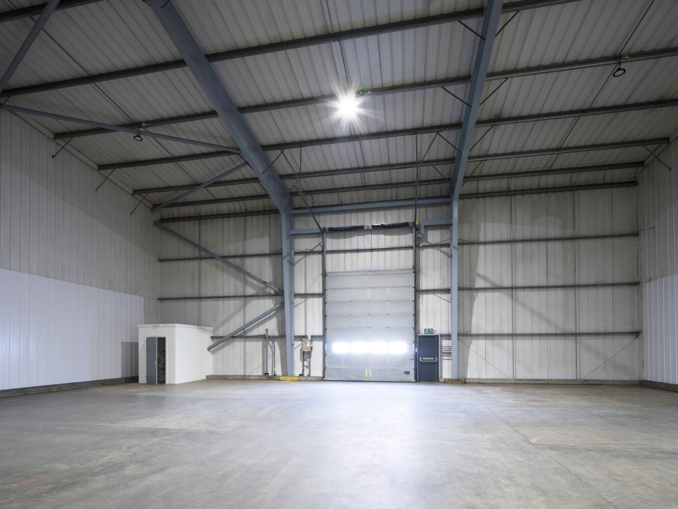 Spacious, empty industrial warehouse with high metal ceiling, concrete floor, large closed overhead door, and small white utility room in the corner.