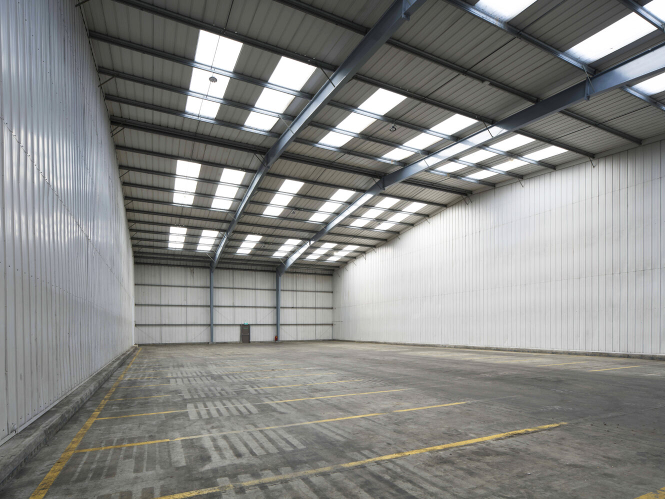 Interior of a large empty warehouse with high ceilings, white metal walls, and skylights. The concrete floor has marked parking spaces and is free of any objects or equipment.