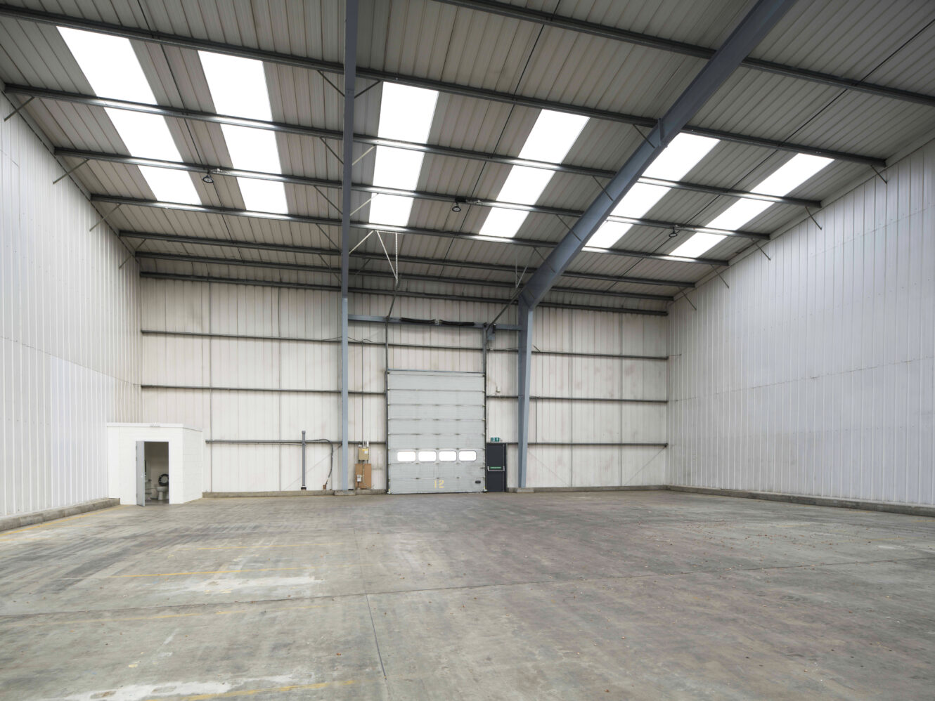 Spacious, empty warehouse interior with high ceilings, skylights, concrete floor, and a large industrial door at the far end. A small white room is in the left corner.