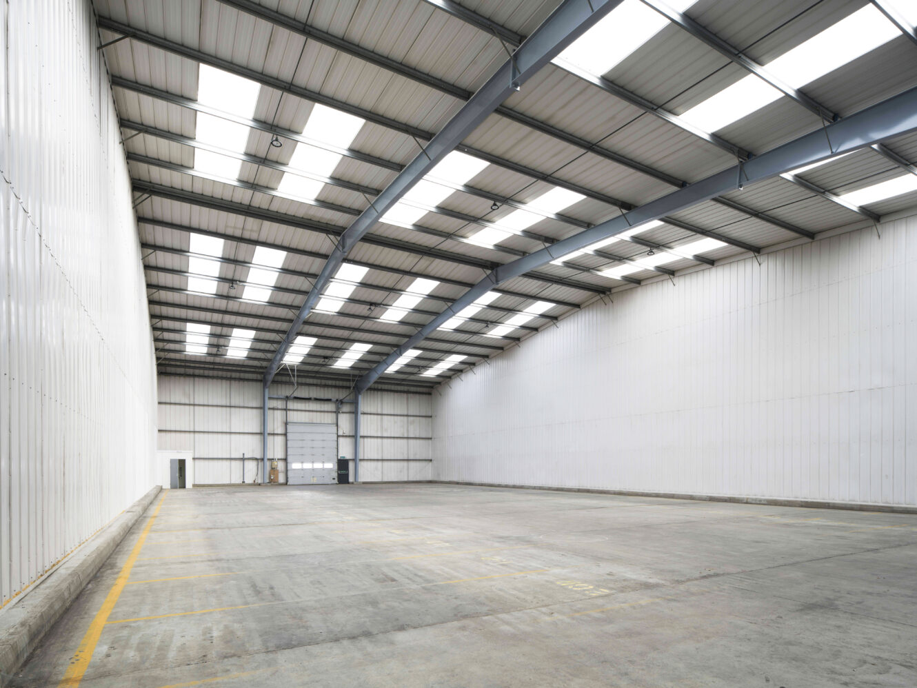 Interior view of an empty warehouse with white walls, high ceilings, skylights, and marked parking spaces on the concrete floor.