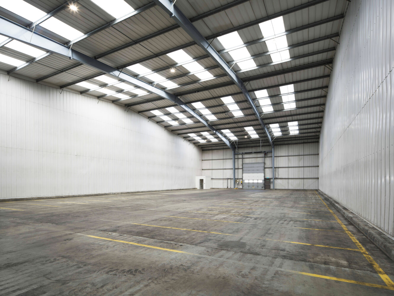 Large, empty warehouse interior with high ceilings, metal beams, skylights, white walls, and a concrete floor marked with yellow lines.