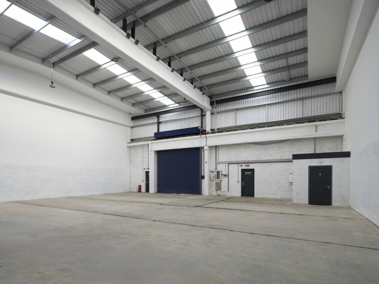 Empty industrial warehouse with a concrete floor, high ceiling, white walls, large blue shutter door, and two smaller black doors on the right. Overhead lights and skylights provide illumination.