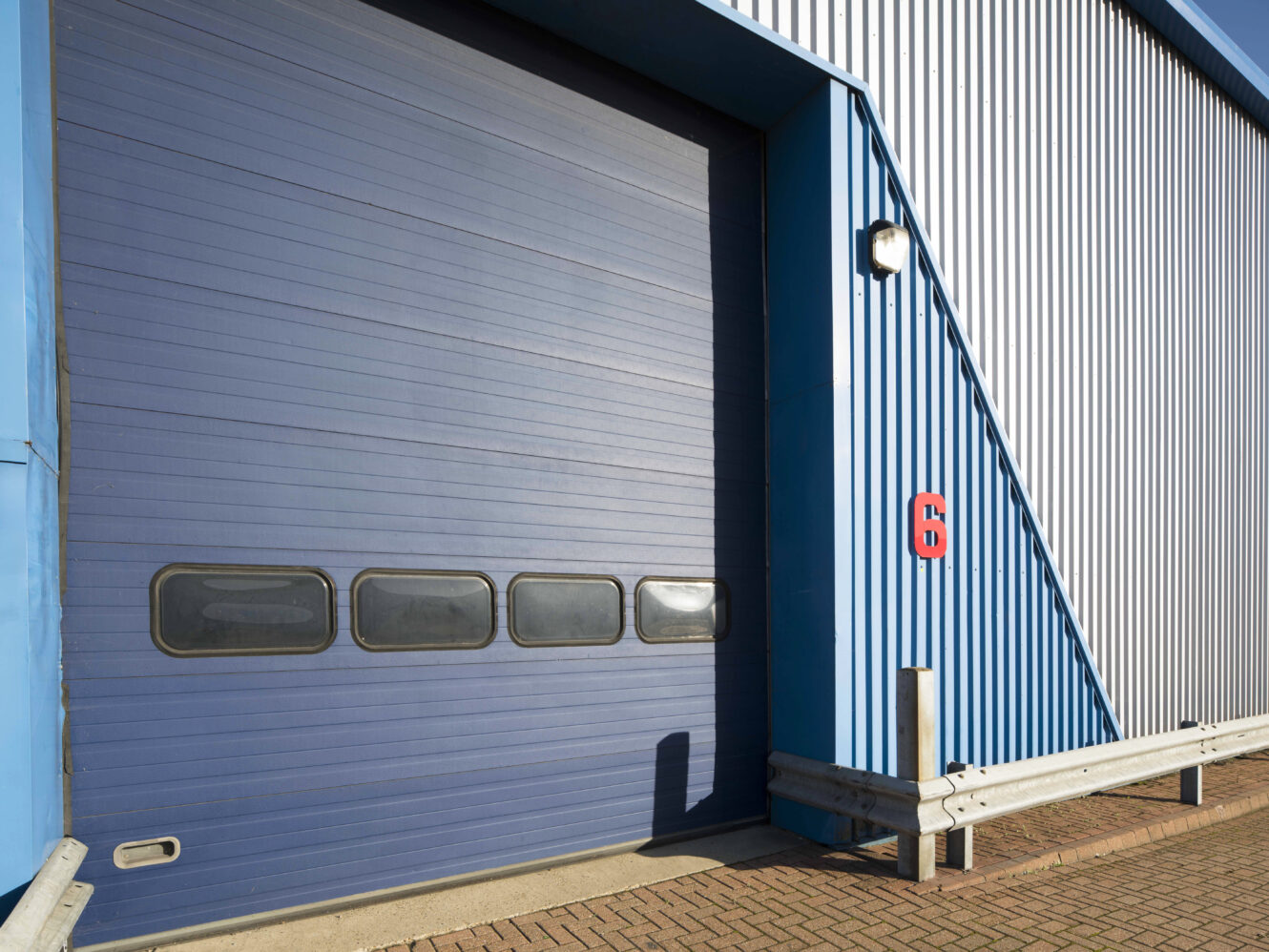 Large blue industrial garage door with four small windows, attached to a corrugated metal building with a red emergency button, under sunlight.
