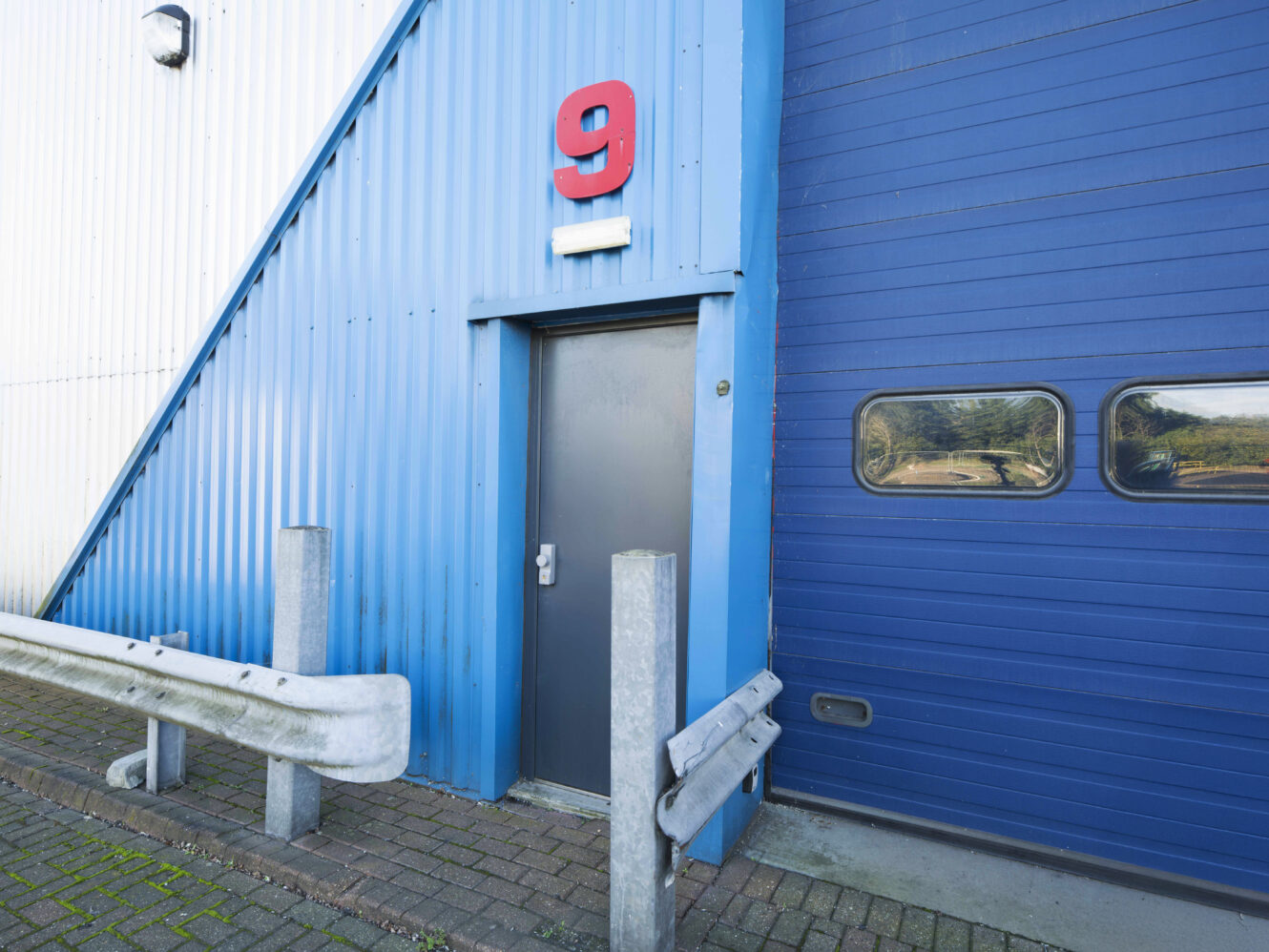 A small gray metal door under a large red number 9 sign on a blue corrugated metal building, with bollards and a guardrail in front.