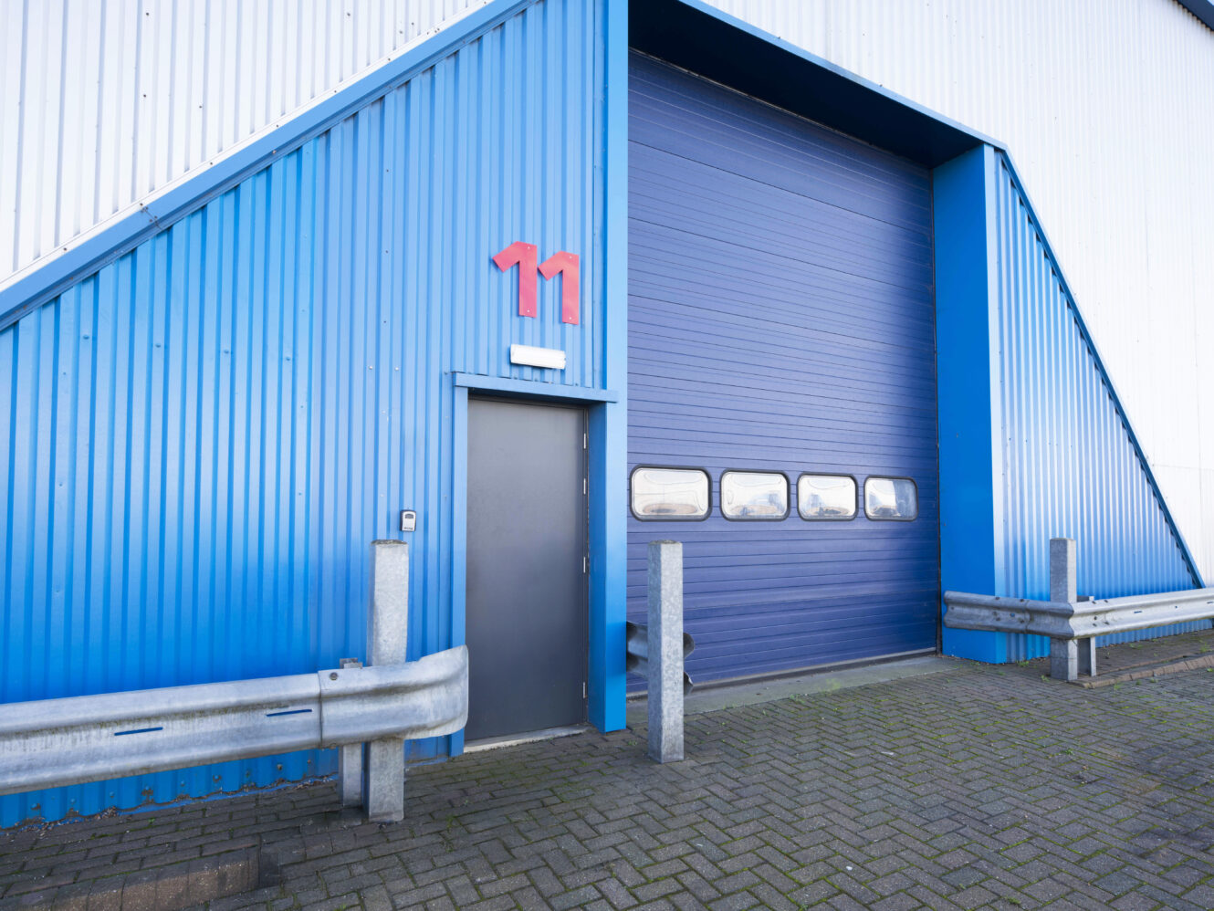 Blue industrial building exterior with a large blue garage door, a small gray door, and a red number 11 above the entrance. Metal barriers and brick pavement are in front.