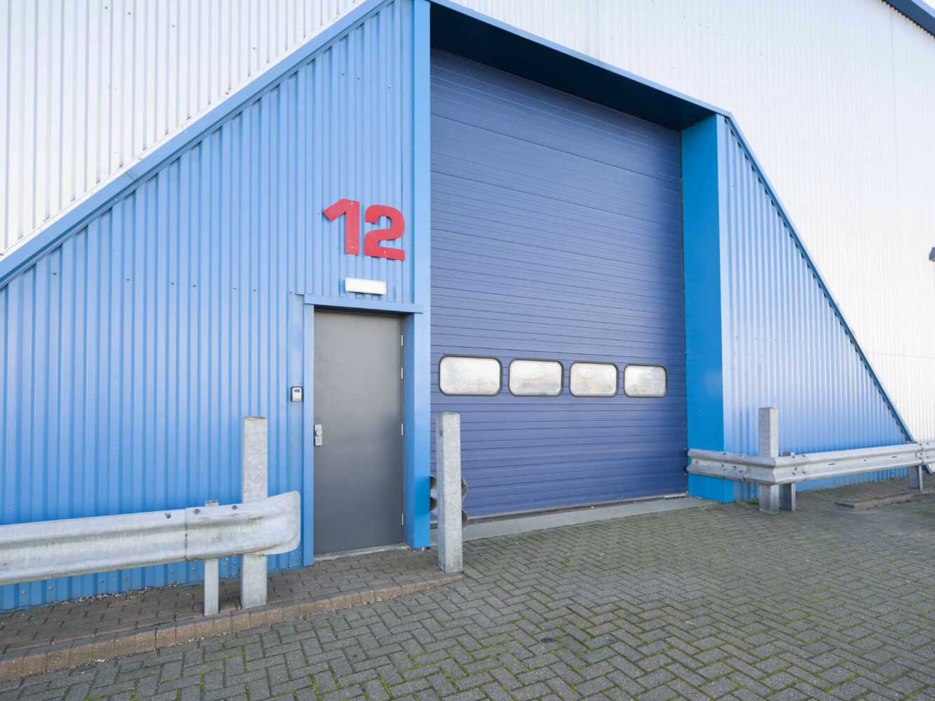 A blue industrial building with a metal door, a large roller shutter, and the number 12 in red above the door. Brick pavement and metal barriers are in front.