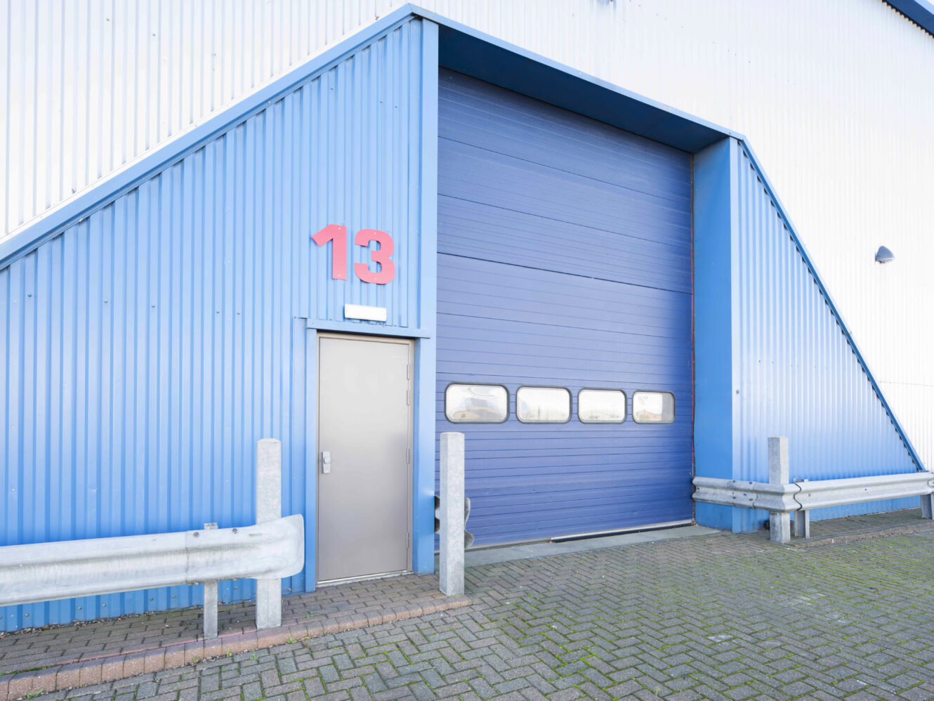 Large industrial blue garage door and a small gray entry door, both set in a corrugated metal building with the number 13 above the entry door.