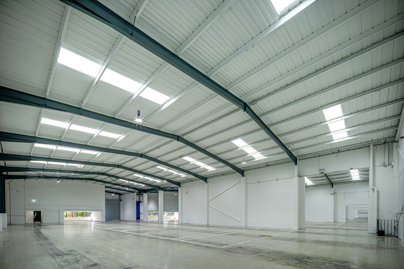 Interior view of a large, empty warehouse with a high, white metal ceiling, skylights, and clean concrete floors.