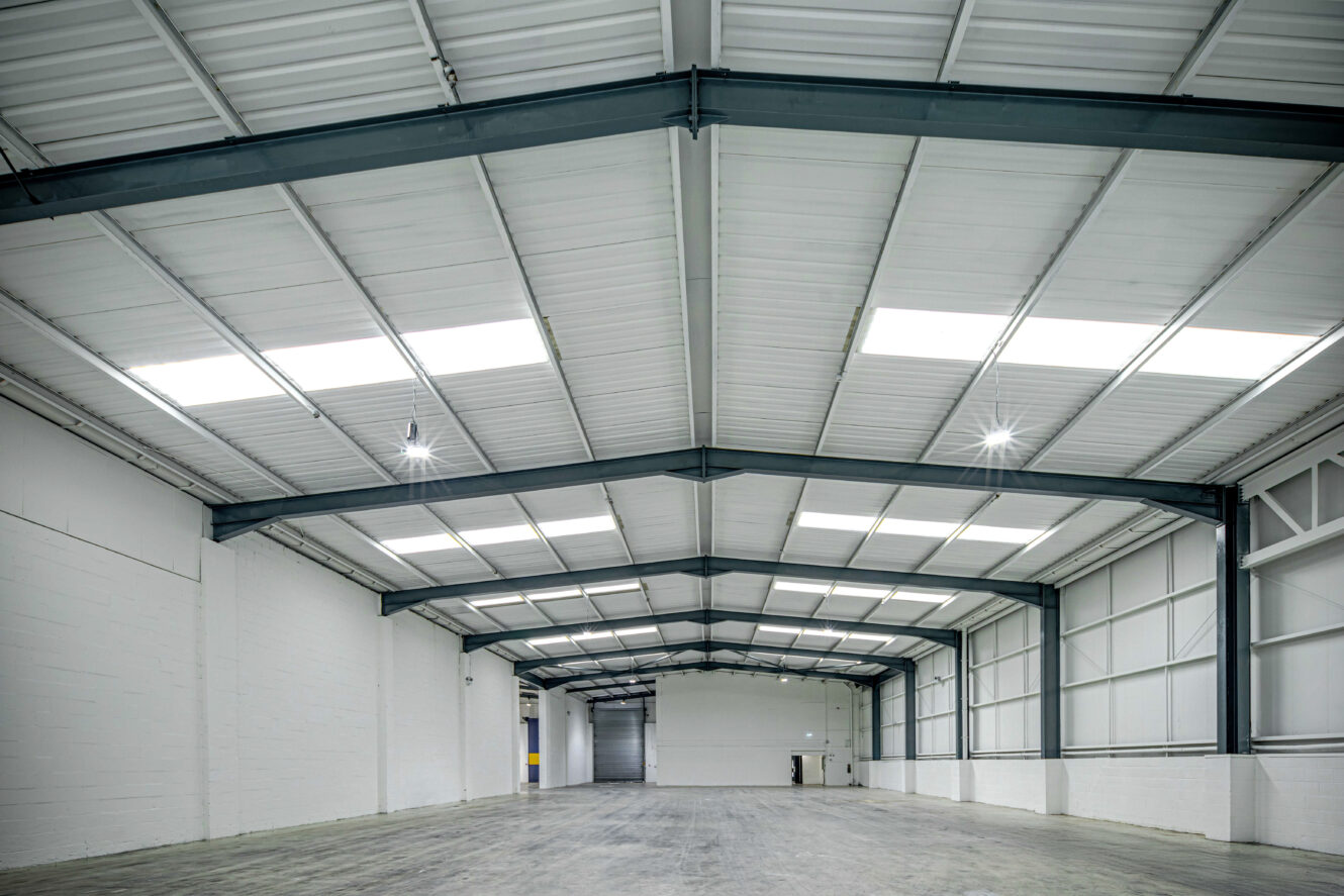 Interior of an empty industrial warehouse with white walls, high ceiling, exposed steel beams, and bright overhead lighting.