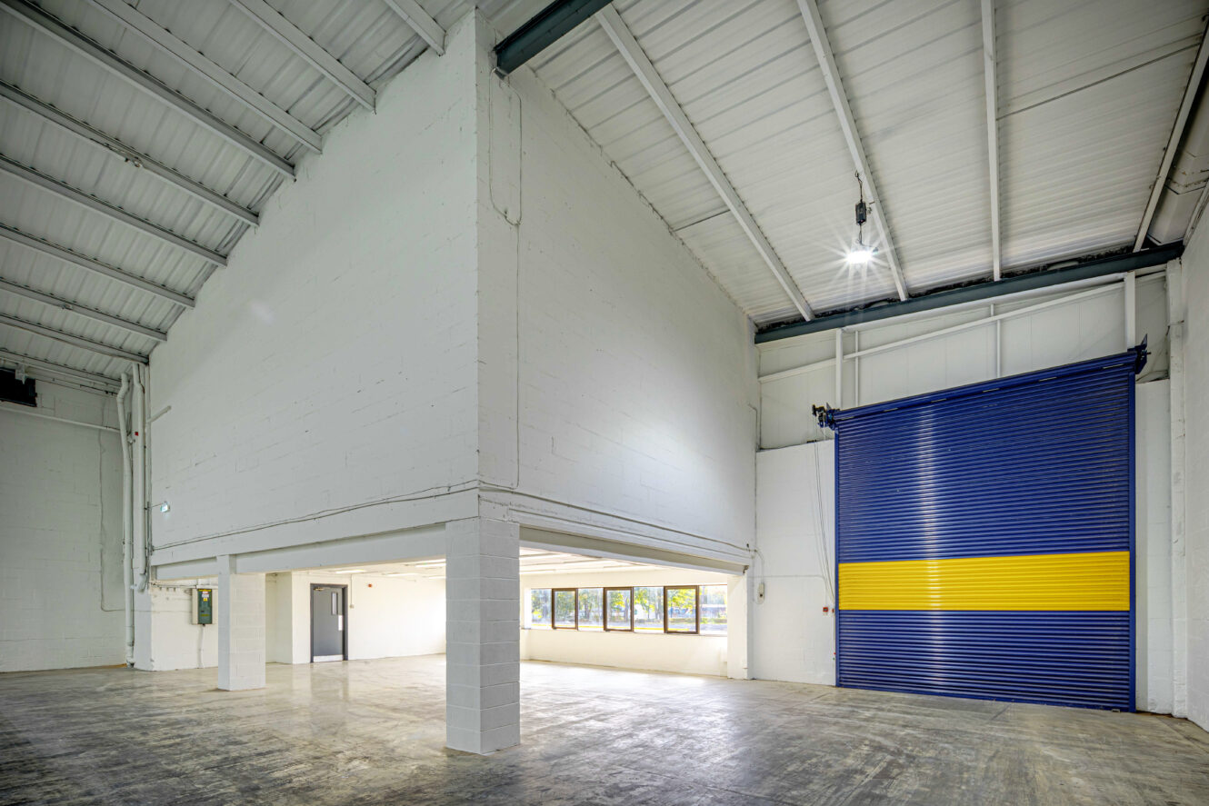 Interior of an empty industrial warehouse with white walls, high ceilings, a mezzanine level, large windows, and a blue and yellow roller door.