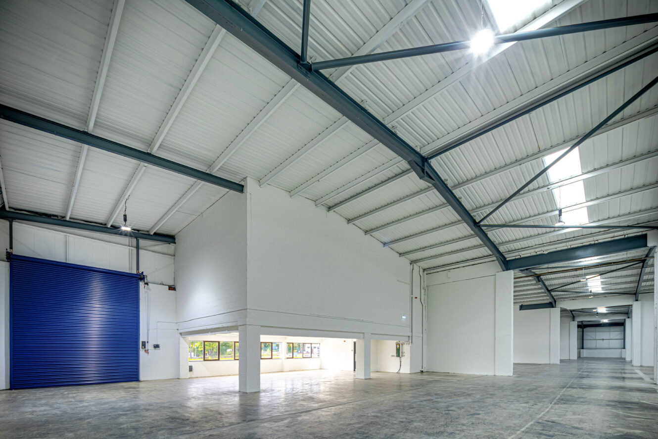 Interior of a large, empty warehouse with high ceilings, exposed steel beams, white walls, concrete floor, and a blue roll-up door.