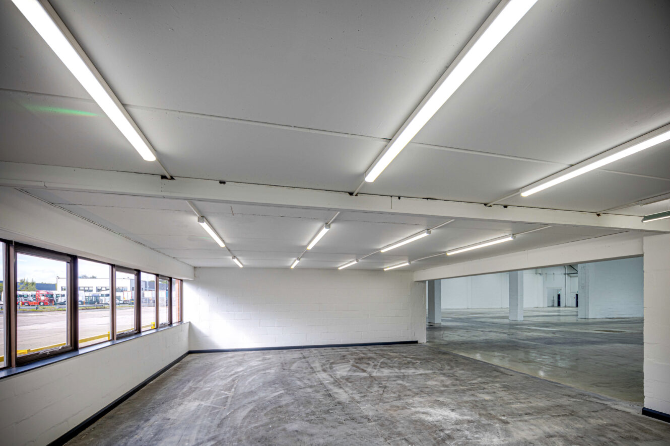 An empty, well-lit industrial space with white walls, fluorescent ceiling lights, concrete floors, and large windows along one side.