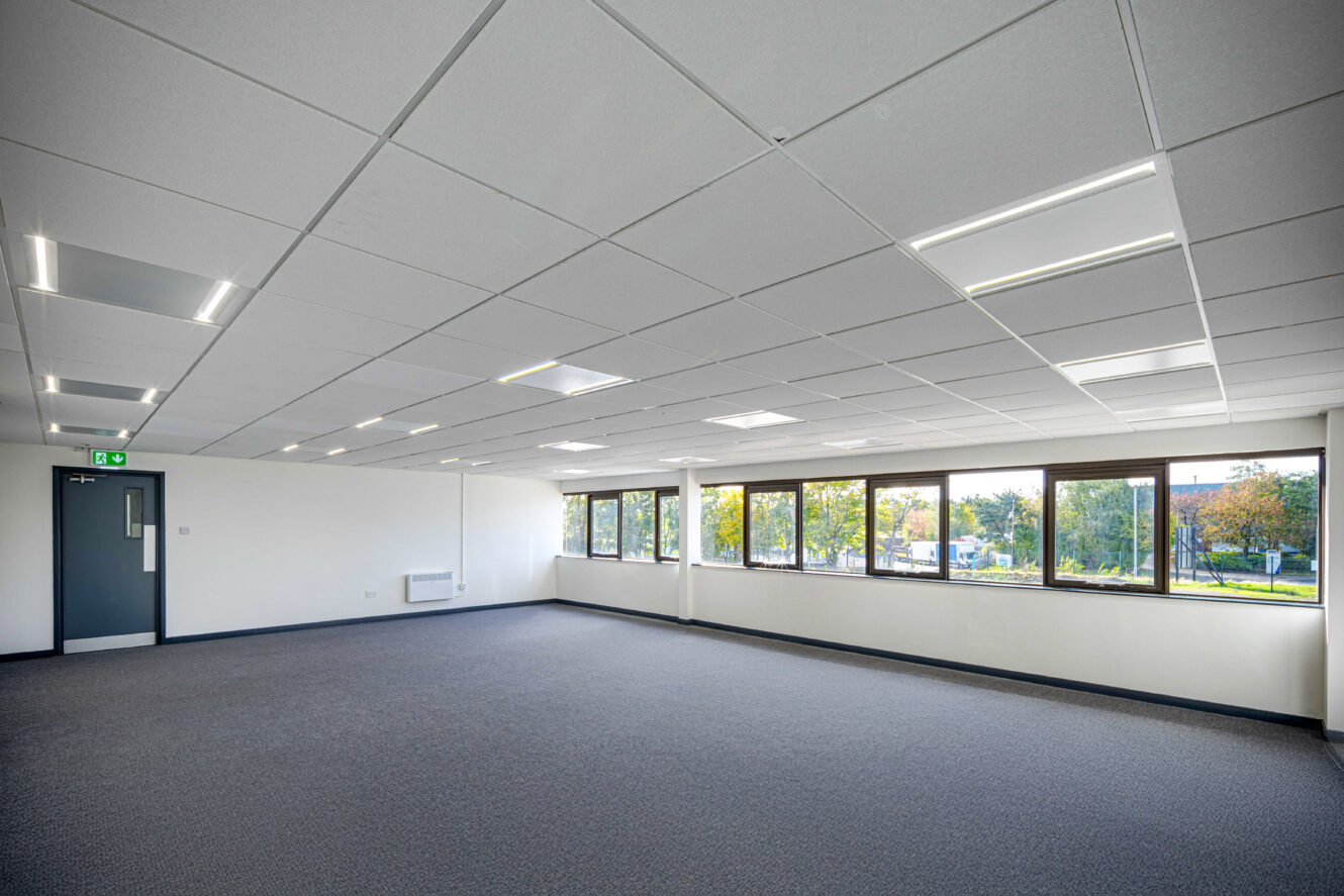 Spacious, empty office room with gray carpet, white walls, large windows letting in natural light, and a closed door on the left side. Ceiling lights are turned on.