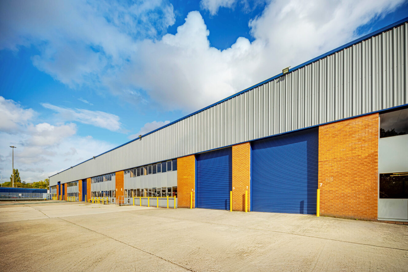 Exterior view of a modern industrial warehouse with brick walls, blue metal cladding, and several large blue roller shutter doors under a partly cloudy sky.