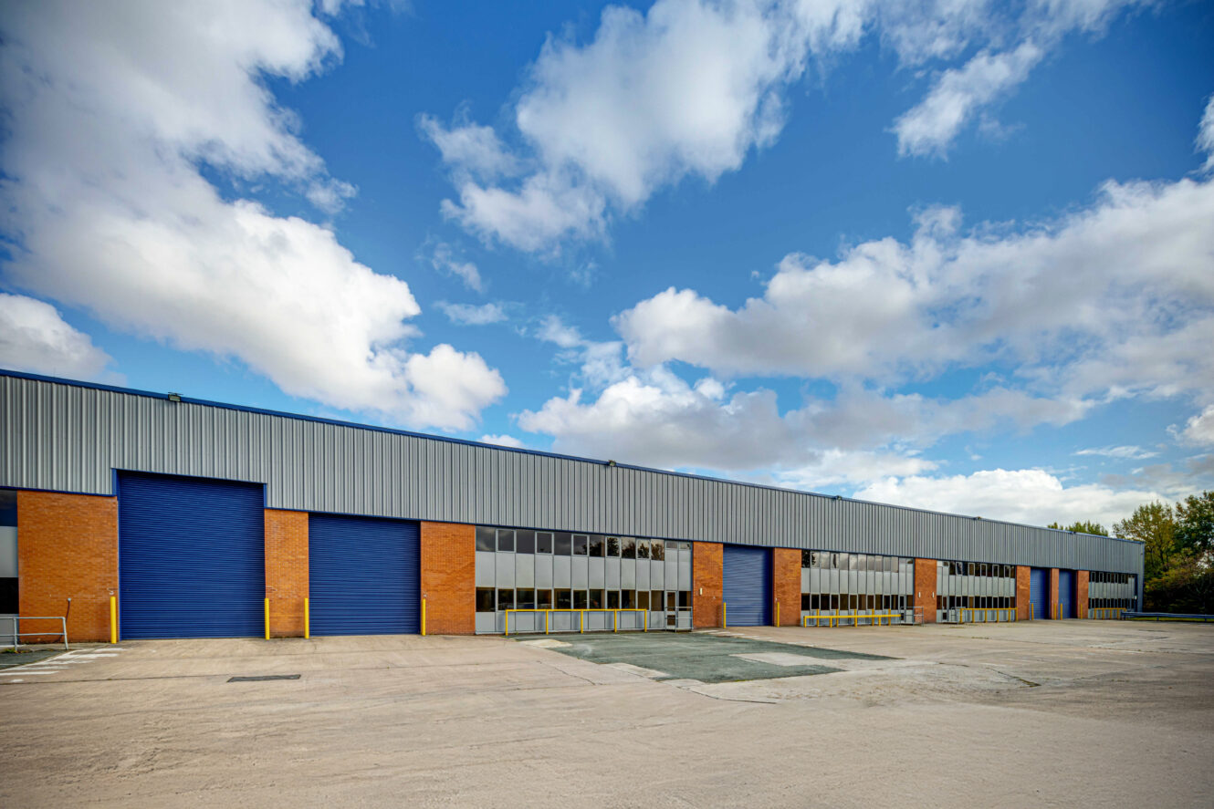 Large industrial warehouse with blue roller doors, brick and metal exterior, and empty paved lot under a partly cloudy sky.