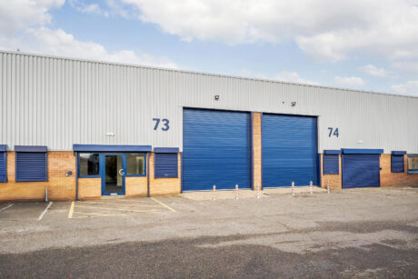 Industrial warehouse building with blue doors and windows, labeled units 73 and 74. The area in front is paved with marked parking spaces and empty bollards.