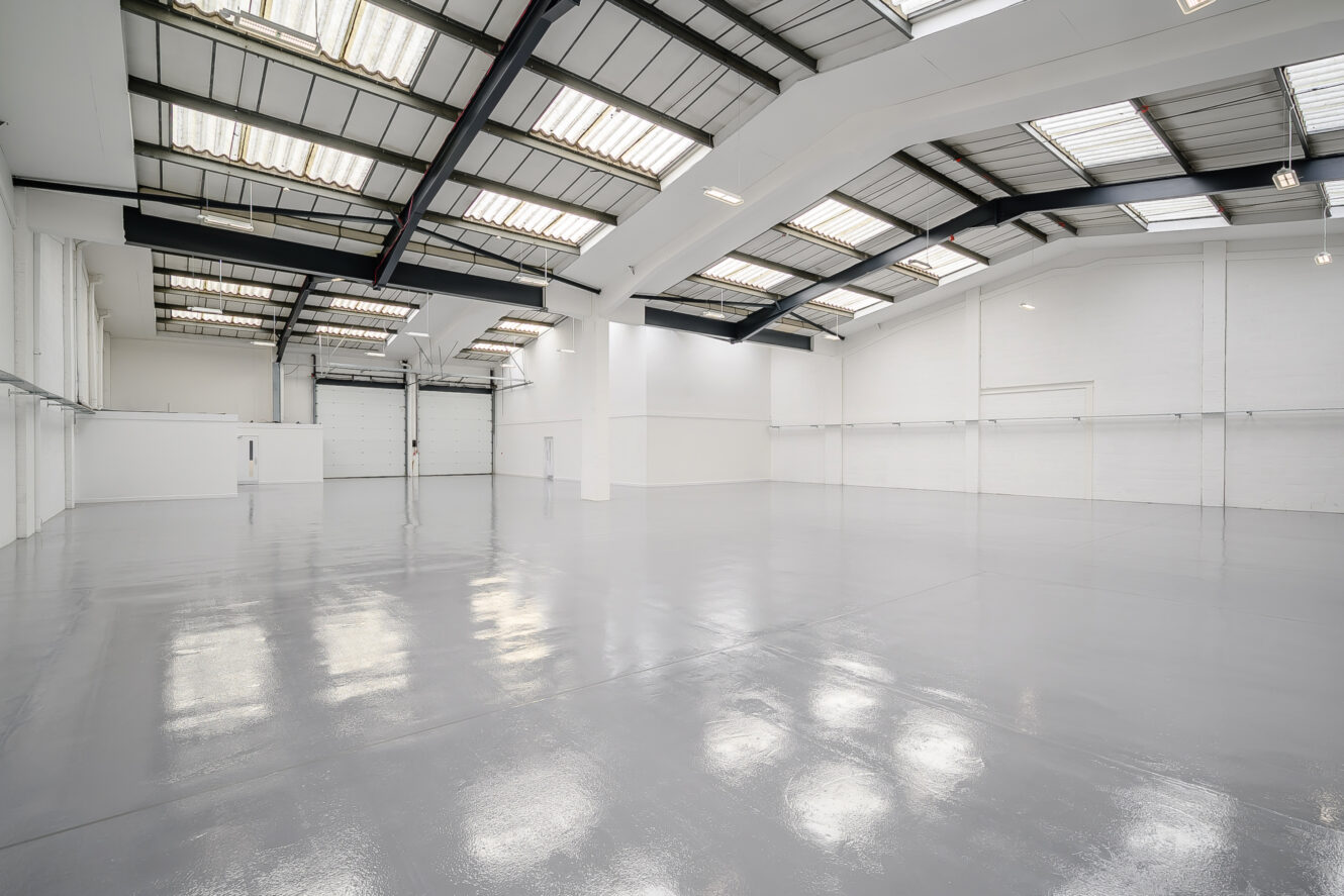 Large empty warehouse with polished concrete floors, white walls, high ceiling, skylights, and industrial lighting.