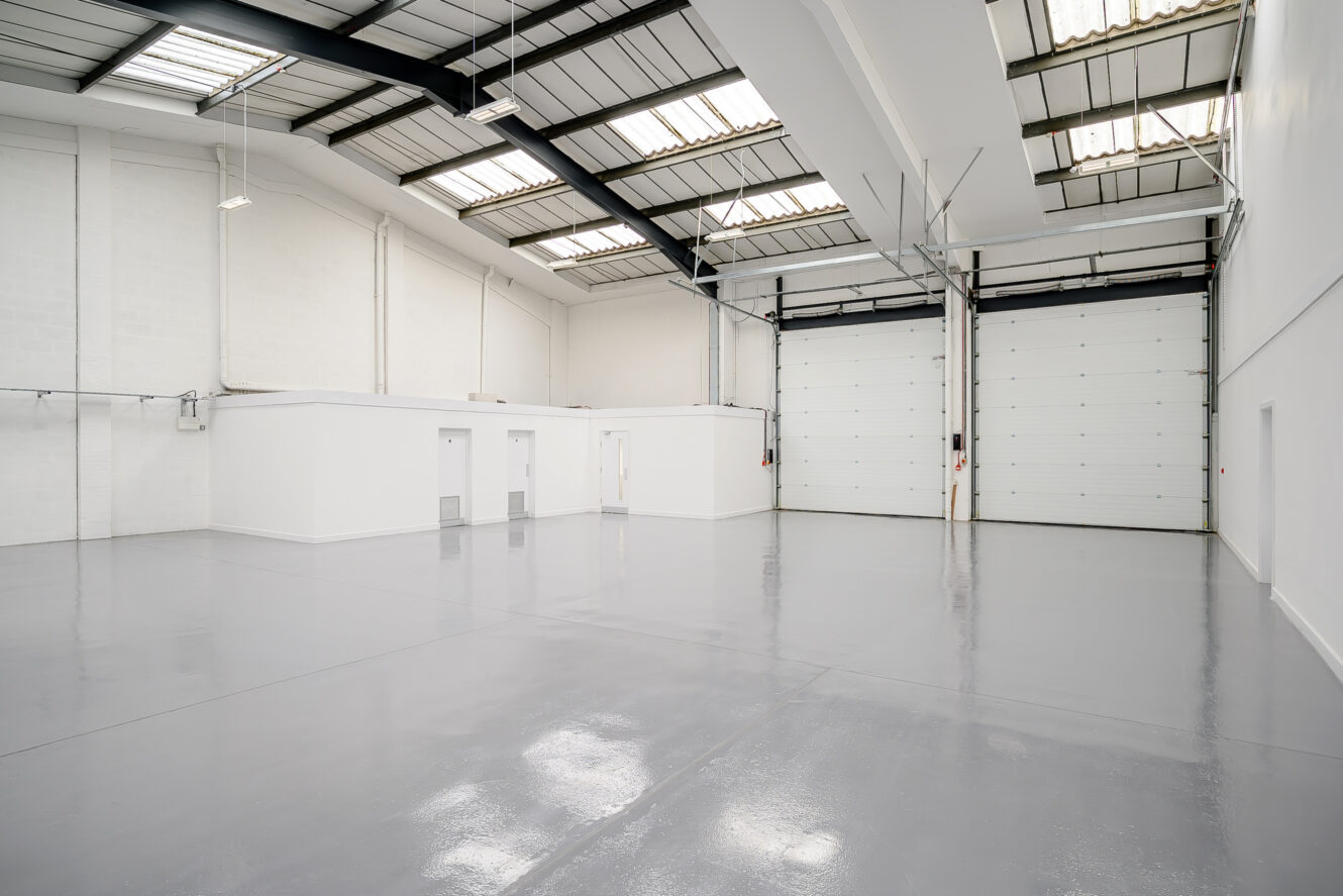 Empty industrial warehouse with glossy gray floors, white walls, high ceilings, and large roller shutter doors; small rooms along one wall.