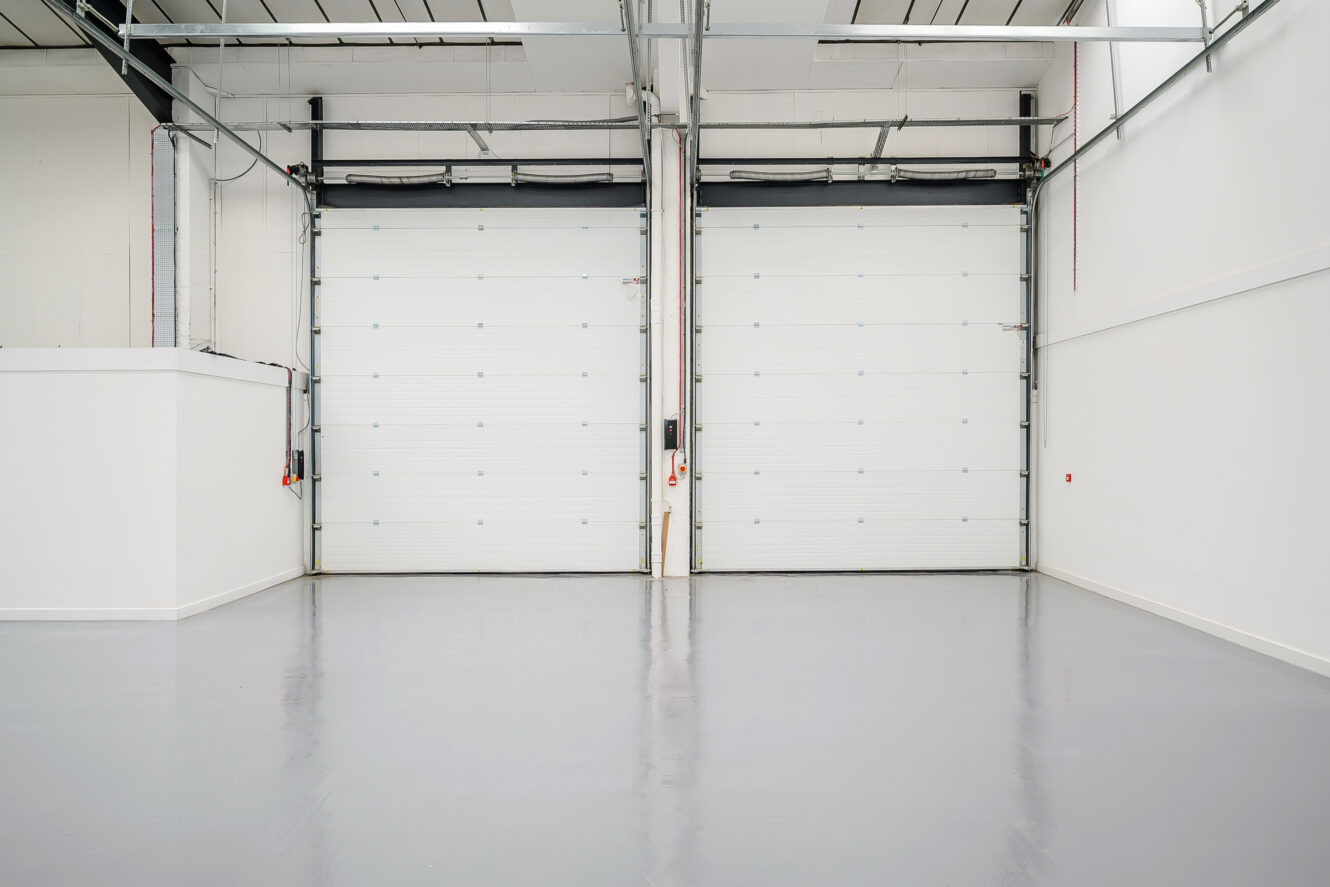 Two large white industrial garage doors in a clean, empty warehouse space with white walls and a smooth gray floor.