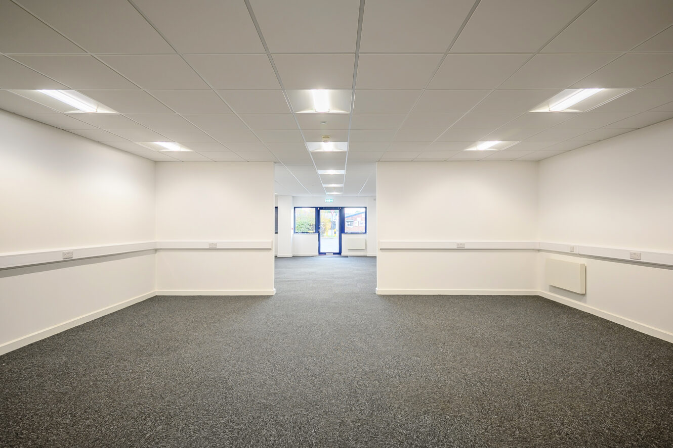 An empty office space with white walls, carpeted floor, ceiling lights, and windows at the far end letting in natural light.