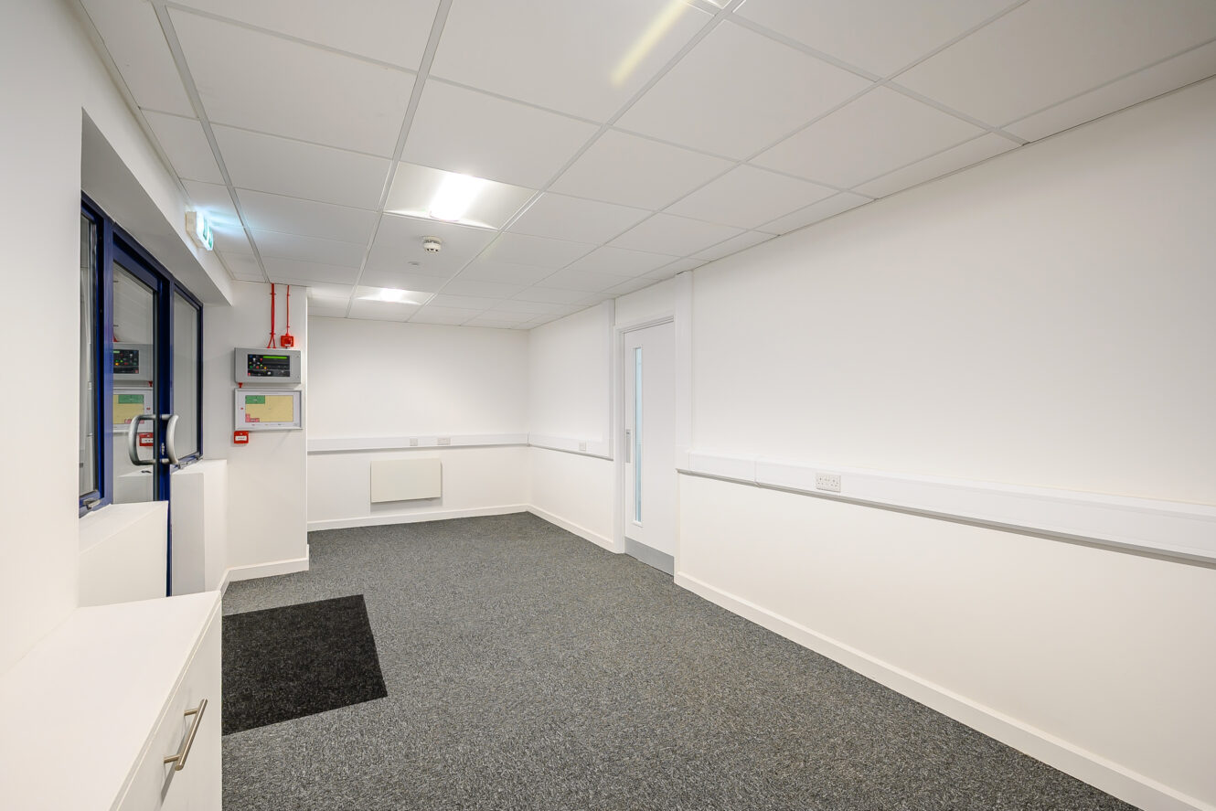 A clean, empty office room with white walls, grey carpet, fluorescent ceiling lights, and a windowed door. Electrical panels are mounted on the left wall.