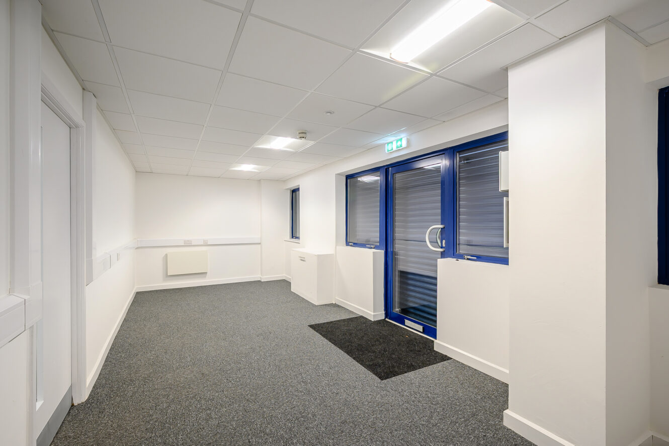 A clean, empty hallway with white walls, grey carpet, and a blue-framed glass exit door under fluorescent ceiling lights.