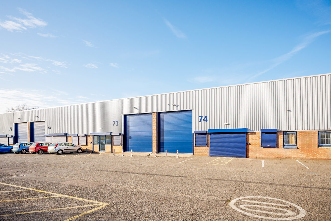 Industrial warehouse building with numbered blue doors 73 and 74, several parked cars, and empty parking spaces on a clear, sunny day.
