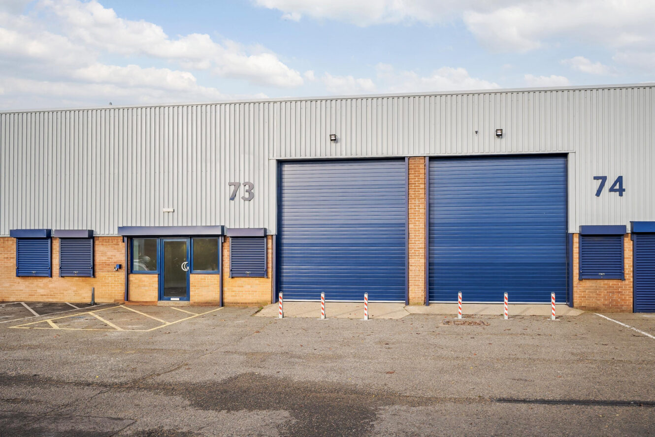 Industrial warehouse exterior with two large blue roller doors, labeled 73 and 74, and a glass entrance door. There are bollards and disabled parking in front.