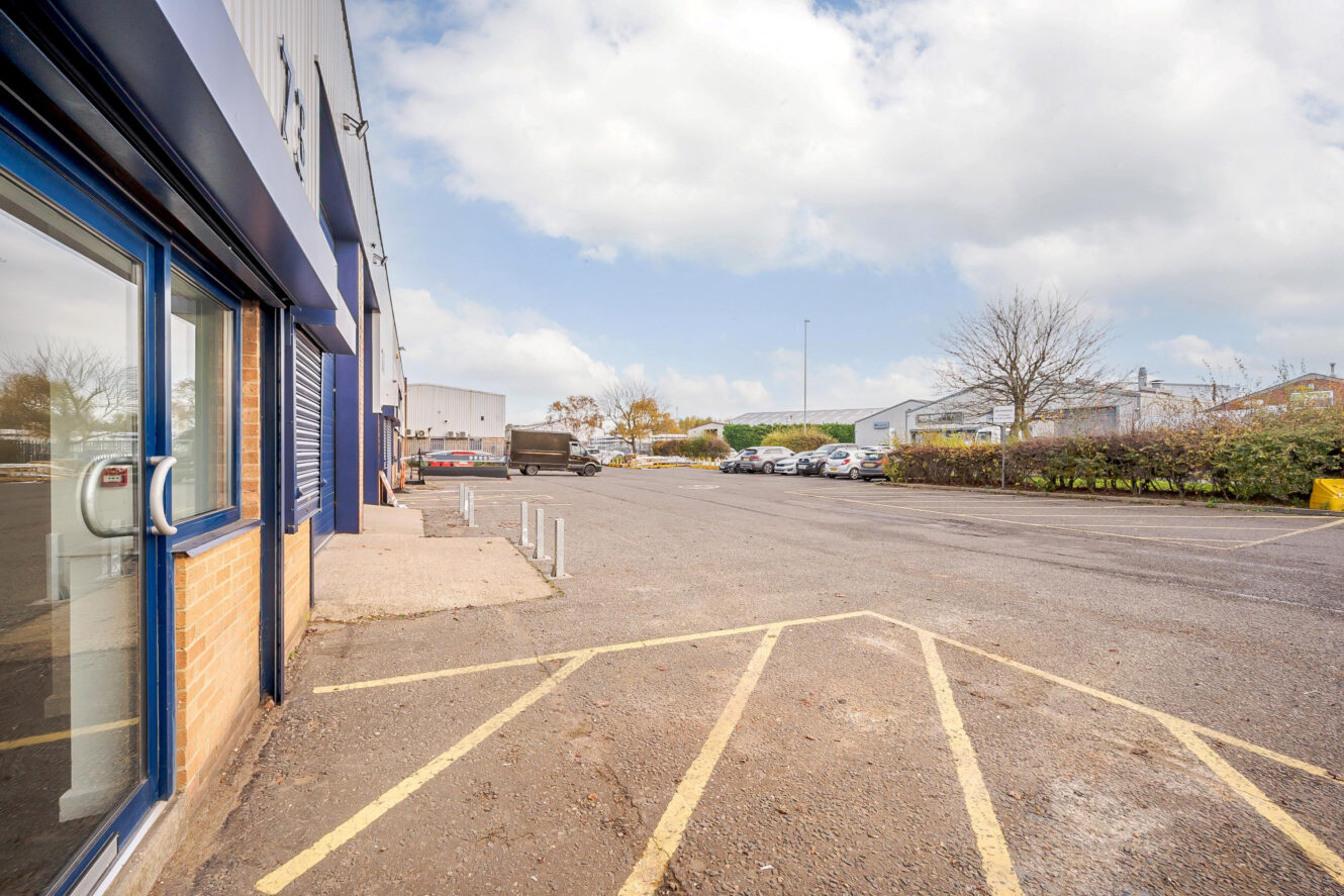 A mostly empty industrial parking lot with yellow painted lines, some parked vehicles, and buildings along the left side under a cloudy sky.