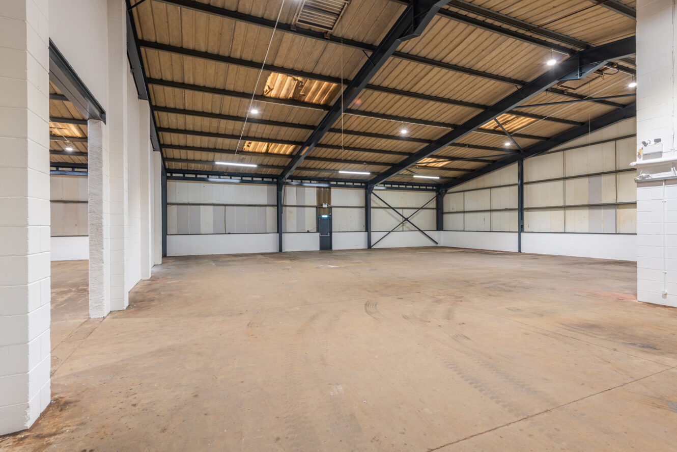 Interior of an empty industrial warehouse with concrete floor, white brick walls, metal beams, and a single door at the far end.