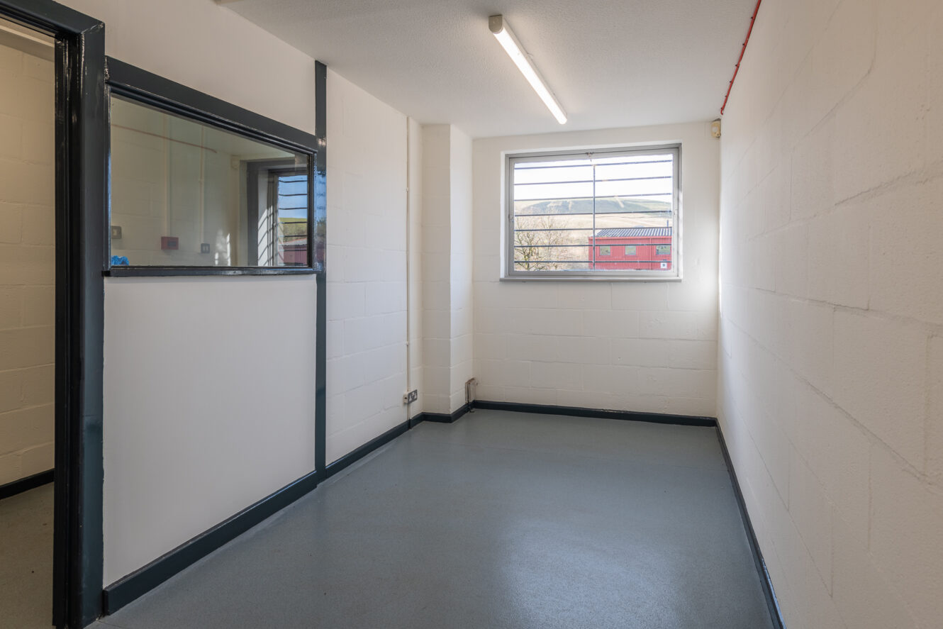 A small, empty office room with white walls, a single window with bars, a fluorescent ceiling light, and a glass partition on one side.
