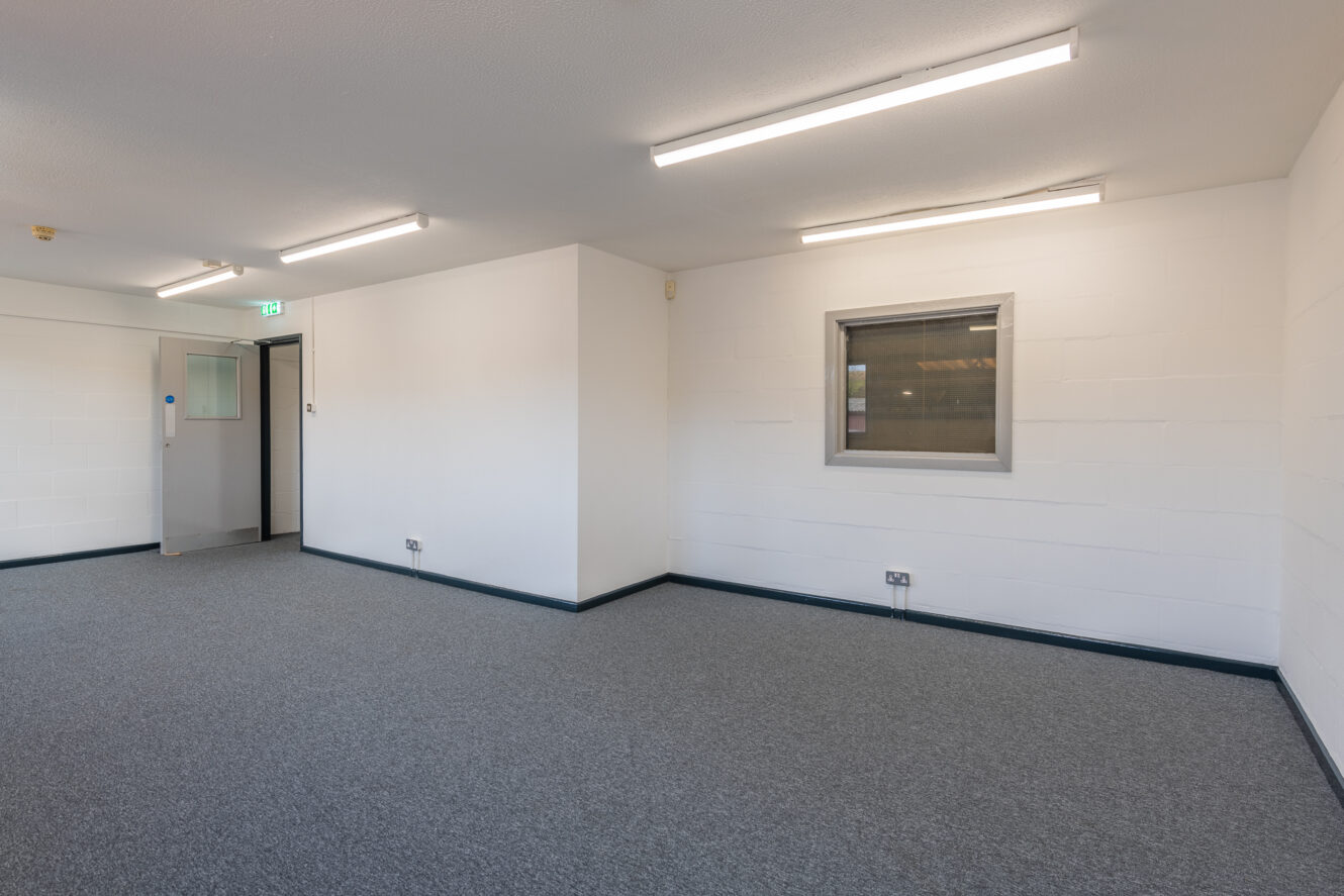 Empty office room with white walls, grey carpeted floor, fluorescent ceiling lights, a window in one wall, and an open door leading to a corridor.