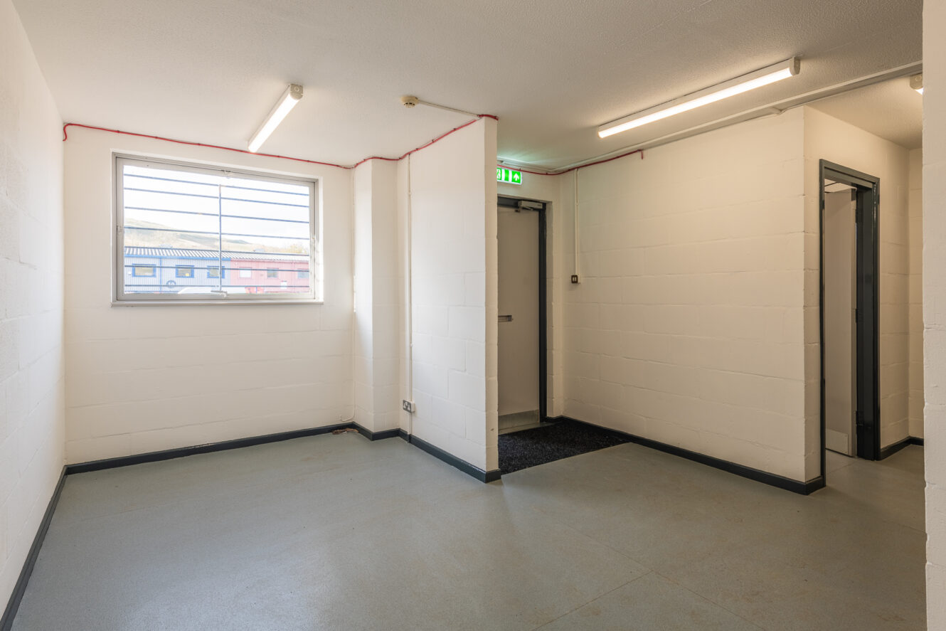 Small, empty room with white walls, a barred window, fluorescent lights, and two doorways; one door has an illuminated exit sign above it.