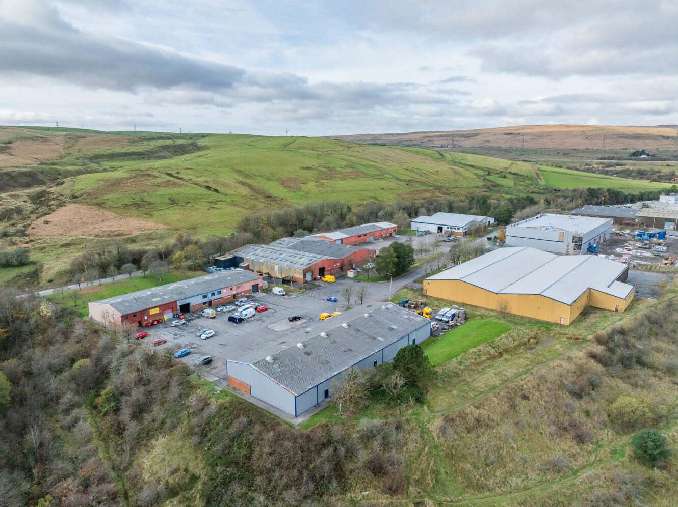 Aerial view of an industrial estate with multiple warehouses, parked vehicles, and surrounding green hills under a cloudy sky.