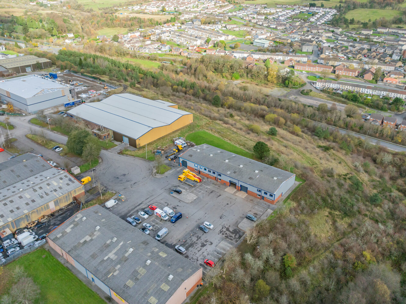 Aerial view of industrial buildings with parked vehicles in a business park, surrounded by greenery and a nearby residential area.