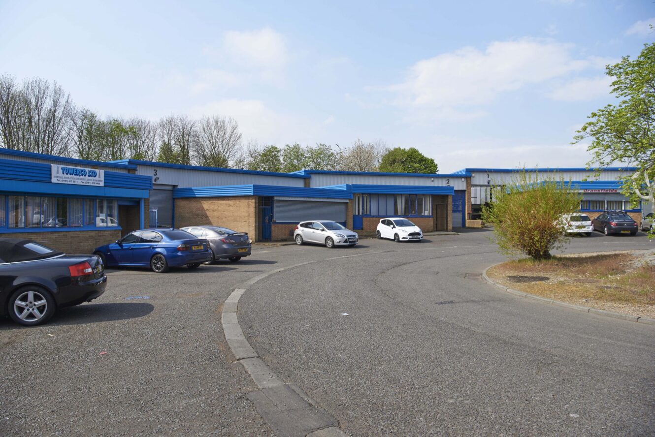A curved driveway in front of several low, blue-roofed commercial buildings with parked cars and trees in the background.
