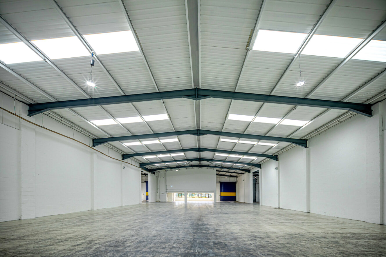 Empty industrial warehouse with white walls, high ceilings, exposed steel beams, and skylights allowing natural light to fill the large open space.