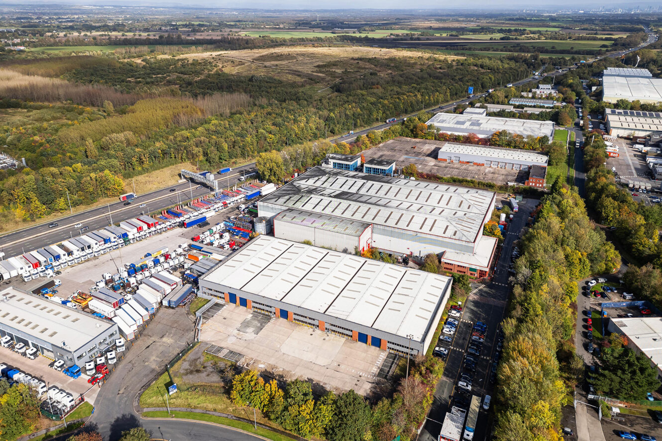 Aerial view of an industrial park with warehouses, trucks, and trailers, surrounded by trees and fields, with a road running alongside the site.