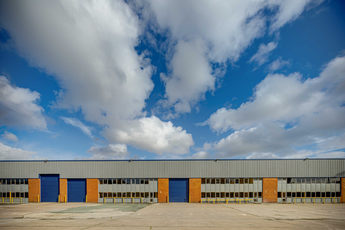 Large industrial warehouse with brick and metal exterior, featuring several blue roller doors, wide concrete forecourt, and a cloudy sky overhead.