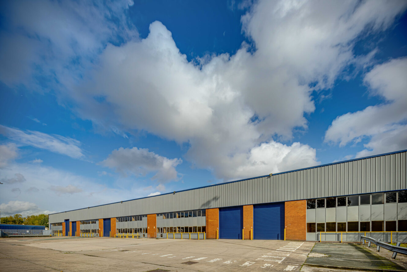 A large industrial warehouse with blue roller doors and red brick walls sits under a partly cloudy sky, with an empty parking lot in the foreground.