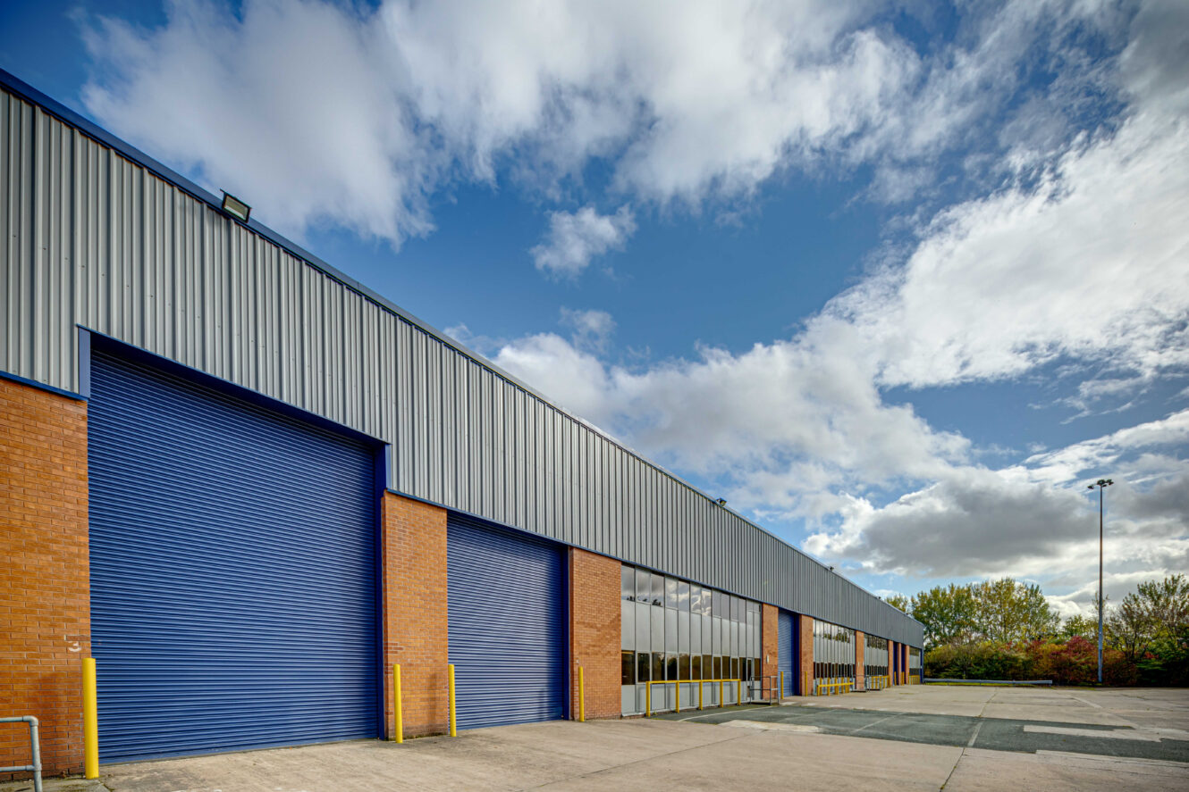 Large warehouse with brick and metal exterior, featuring several blue roll-up doors and a wide driveway under a partly cloudy sky.